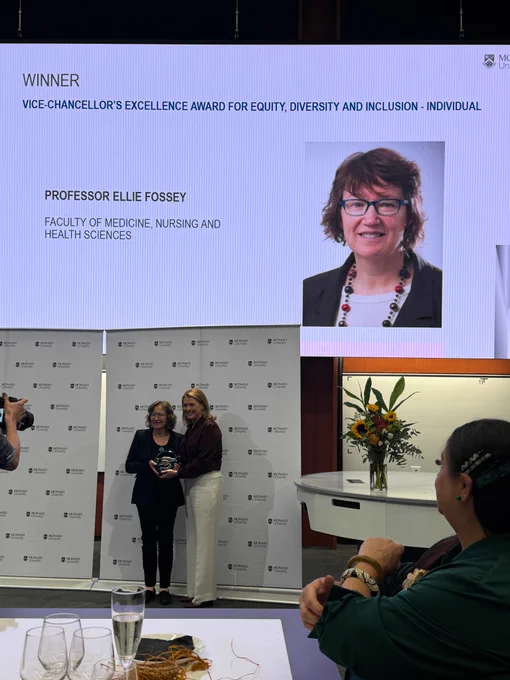 Two people standing holding a trophy below a large data projector screen with an image of a person in it which says "Winner: Vice-chancellors excellence award for equity, diversity and inclusion - individual" and has the name Professor Ellie Fossey below it