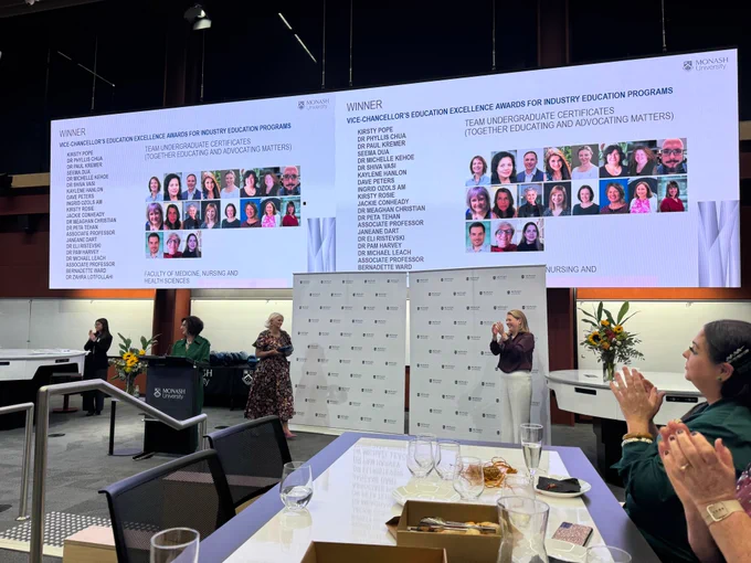 A group of people clapping with a data projector dual screen showing headshots of a large group with the heading "Winner: Vice Chancellors Education Excellence Award for Innovation in Education Programs"