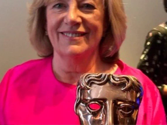 Dianne Nelmes holding a BAFTA award, wearing a bright pink top and smiling at the camera.