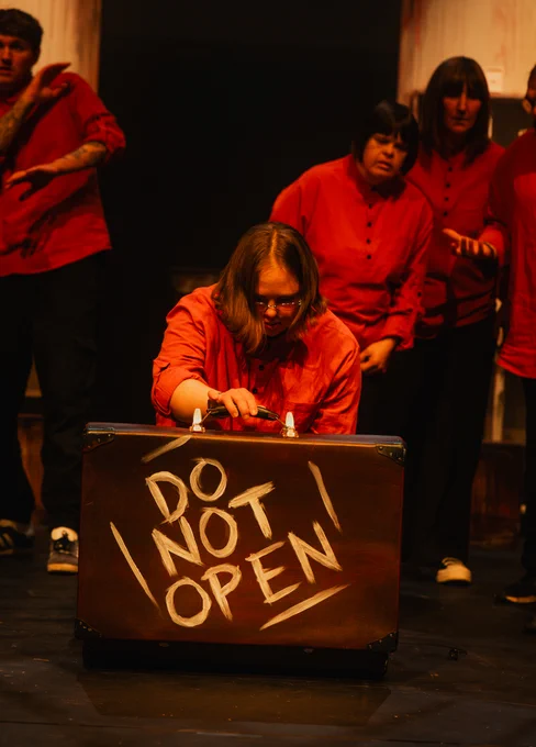 A performer in a red top kneeling and holding the handle of a suitcase that says DO NOT OPEN in large letters on it. Other performers in the background are standing and watching her.