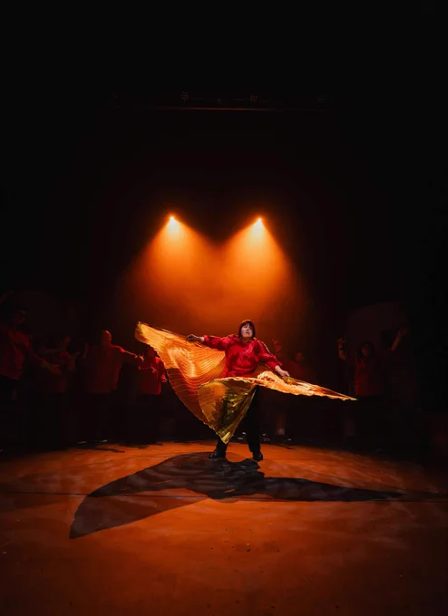 A performer in a red top and black trousers, spot lit as she swirls gold wings around her.