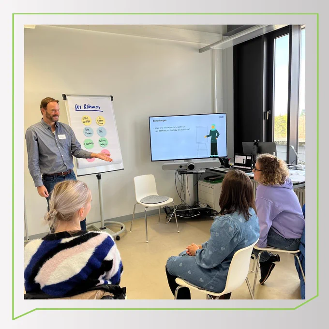 Tobias Maier steht vor einer Flipchart und einem Bildschirm. Auf dem Bildschirm ist ein Avatar vom E-Learning zu sehen. Vor ihm sitzen drei Teilnehmerinnen, die insteressiert zuhören. Tobias erklärt etwas.
Hintergrundfarbe: Hellgrauer Verlauf, grüne Umrandung in der Form der NaWik-Sprechblase.