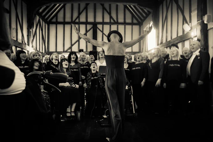 A black-and-white photograph of a choir performance inside a timber-framed hall. The conductor, standing with arms outstretched, faces the choir, which consists of diverse members, some seated and others standing. The choir appears animated and engaged, singing passionately, with the setting creating an intimate and uplifting atmosphere.