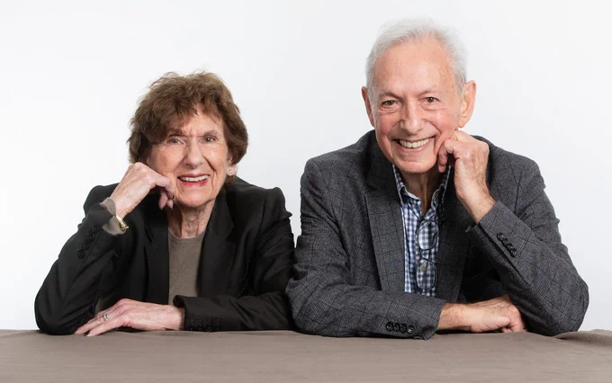 Myrna and Adrian are sat next to eachother smiling at the camera.