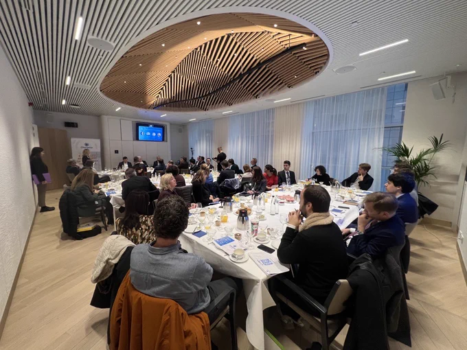 Attendees sit around long breakfast tables in a bright modern room with their attention directed to the four panellists, seated below a projector screen at the end of the room.