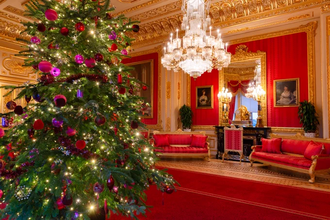 An ornate room with elaborate gold decorations and red walls. A large Christmas tree, adorned with red and gold ornaments, stands prominently on the left, illuminated by festive lights. Elegant crystal chandeliers hang from the ceiling, and luxurious red sofas are arranged around the room. Ornate frames displaying portraits hang on the walls.