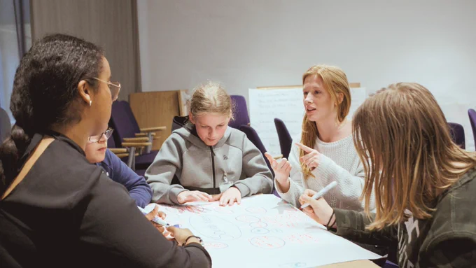 A diverse group of students with and without disabilities sit at a table, writing and listening to a teacher, who is also sitting at the table.