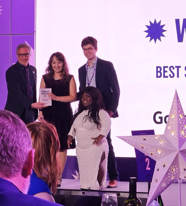 Audience shot of CEO Beth Kume-Holland and Dan Mercer standing on stage with presenter Gavin Lawrie and Fats Timbo, holding the award and posing for the official photograph.  