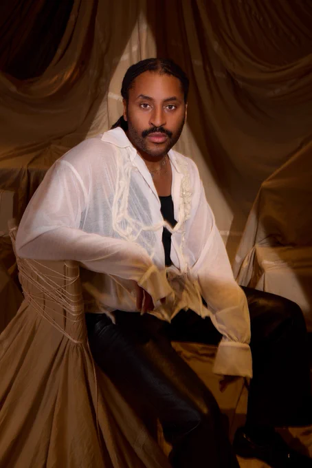 A man with dark hair tied back and a dark moustache. He is wearing a floaty white shirt and black leather trousers.