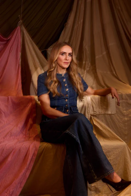 A woman with long blonde curled hair, wearing a denim jumpsuit.