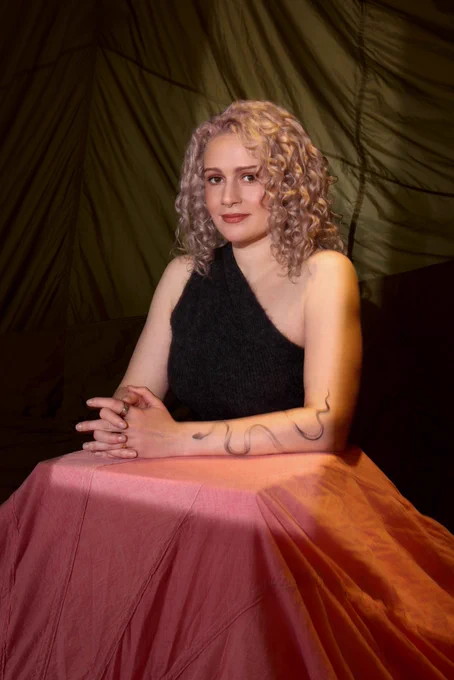 A woman with bobbed dyed grey hair and a snake tattoo on her arm, wearing a black off the shoulder top and white trousers.