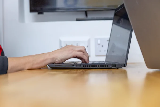 Two hands typing on a laptop. The laptop is on a light brown wooden desk. 