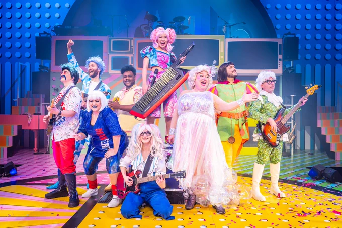 Bright and colourful group photo of the cast of The Rock 'n' Roll Panto Rapunzel, dressed in vibrant costumes. They are on a lively stage set with musical instruments, including a guitar and keytar, and are posing energetically against a playful backdrop featuring vibrant colours. The photographer is Marc Brenner.