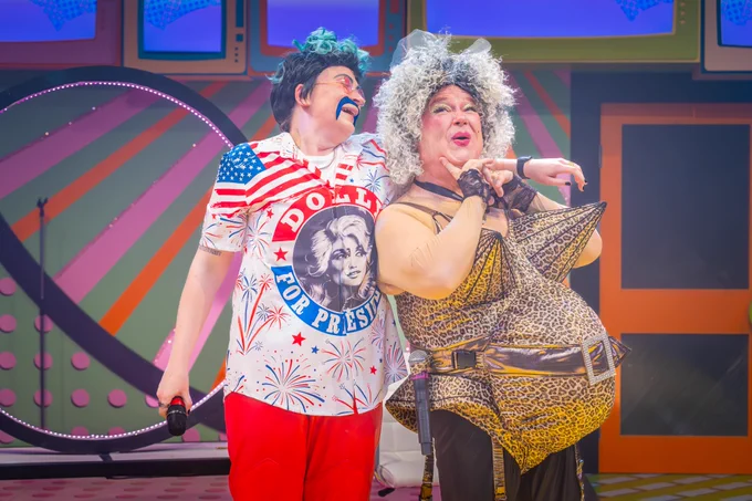 Zoe West as Danny Ruff and Michael Starke as Debbie Updo in The Rock 'n' Roll Panto Rapunzel. Zoe is wearing a bold 'Dolly for President' shirt with red trousers, while Michael is dressed in a leopard-print costume with dramatic accessories. The photographer is Marc Brenner.