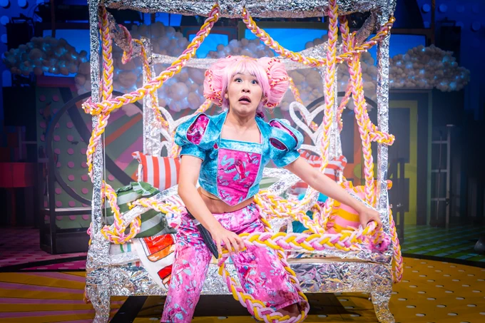 Ai Kumar as Rapunzel in The Rock 'n' Roll Panto Rapunzel, wearing a vibrant pink and blue costume with exaggerated puffed sleeves and styled pink hair. The photographer is Marc Brenner.