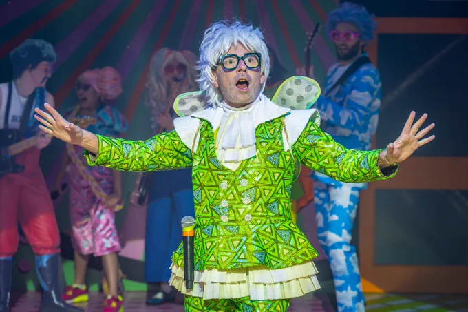 Adam Keast as the Fairy Fixer-Upper in The Rock 'n' Roll Panto Rapunzel, dressed in a bright green, patterned costume with shimmering details and white ruffled accents. The photographer is Marc Brenner.