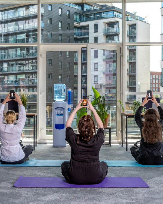Three people raise their phones above their heads on yoga mats wearing office clothes