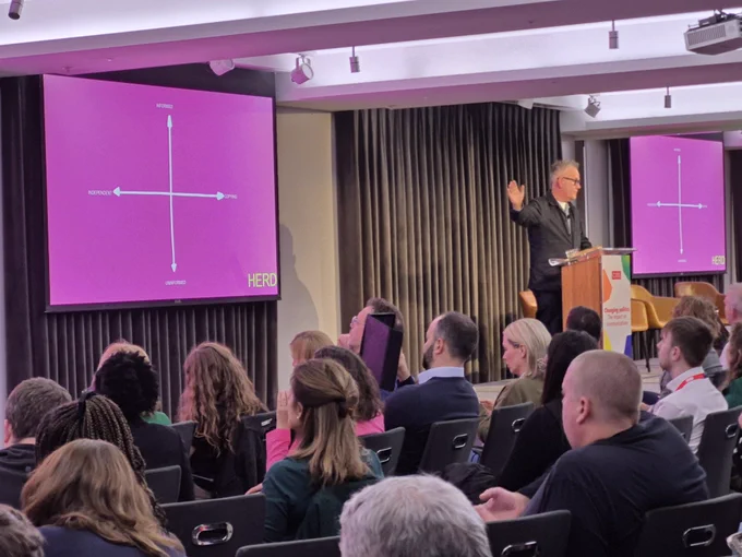 A photo of a man on stage pointing at a pink grid on a slide with four axis.