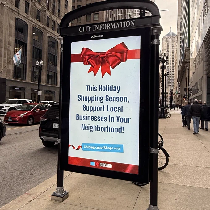 Advertisement on a city street encouraging people to support local businesses during the holiday season, featuring a large red bow and the website 'Chicago.gov/ShopLocal'.