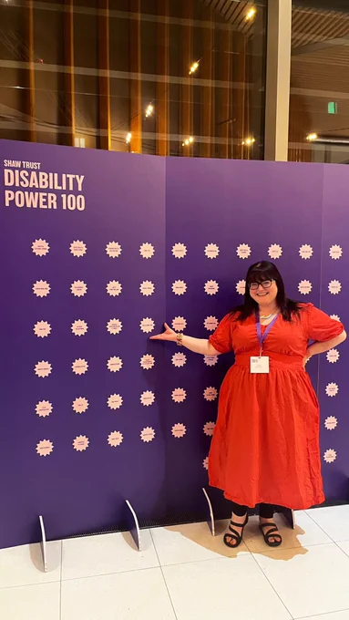 Shaw Trust Disability Power 100; Chella Quint posing in front of a purple wall with white stars that holds the names of awardees. Chella has her hand placed underneath her name.