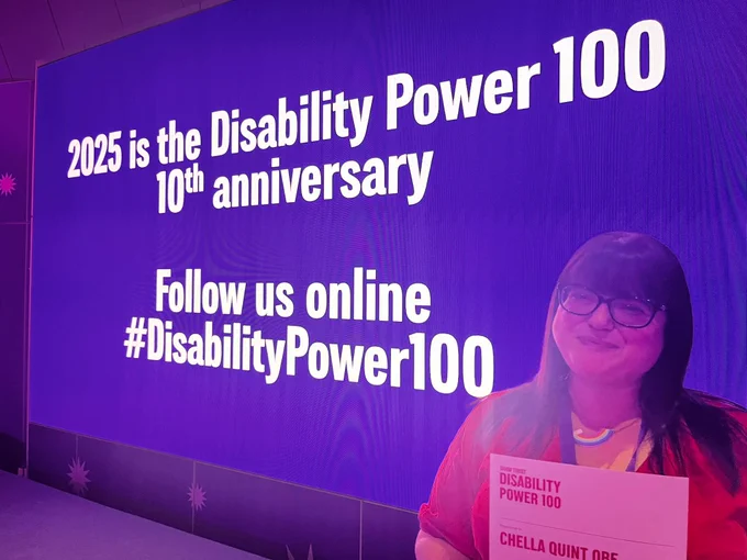 Chella Quint posing with her Disability Power 100 certificate against a blue screen with white text that reads: 2025 is the Disability Power 100’s 10th anniversary. Follow us online #DisabilityPower100.