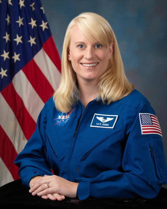 NASA astronaut Kate Rubins, a white woman with blonde hair and gray eyes, poses for an official portrait in front of the United States flag. She wears a blue flight suit that has three patches on it: a NASA meatball, her name, and the American flag. Credit: NASA/Robert Markowitz