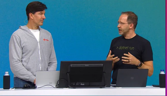 Printscreen from a Microsoft Iginte keynote: Two men (Mark Russinovich on the right, wearing a gray jacket, and Scott Hanselman on the left, wearing a black T-shirt that says "dotnet run") on stage at Microsoft Ignite. On the table are a monitor and a laptop.