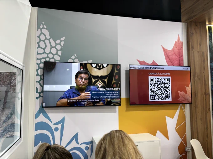 An exhibition booth featuring two screens. The left screen displays a video of Andy Puglas, an Indigenous leader, discussing his community's role in environmental stewardship (in French subtitles). The right screen displays a QR code linked to the "Programme des événements" (event schedule) under Canada's COP29 participation. 