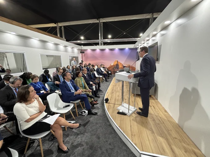 A formal event at COP29, showing a man giving a speech at a podium in a conference space. The audience includes diverse attendees seated and attentively listening, with a scenic backdrop featuring a landscape image. 