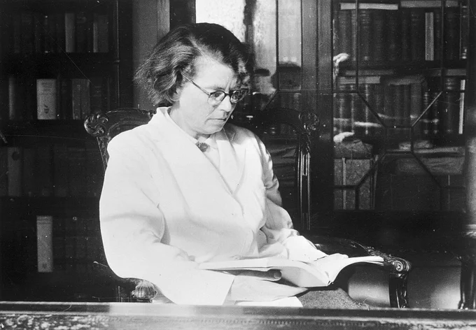 A woman sitting at a desk in a bright white coat, looking engrossed in a book. Behind her is a book cabinet with glass doors.