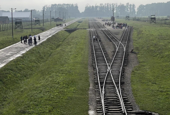 This image captures the rail tracks leading into Auschwitz II-Birkenau site. The mist in the distance adds a haunting, ethereal quality. Visitors walk alongside the tracks.