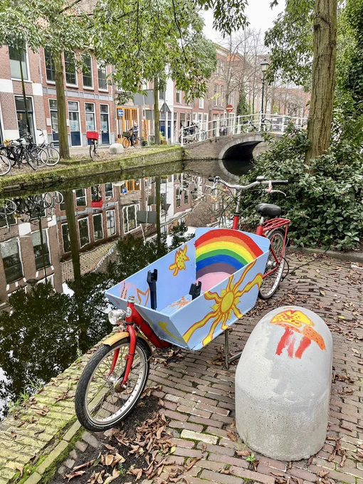 A cargo bike painted with sunshine and rainbows in bright colors is parked next to a canal lined with trees and buildings in Delft.