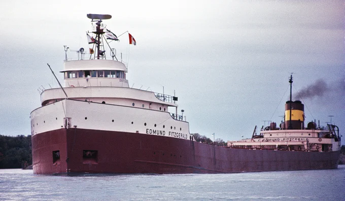 The image shows a large lake freighter ship named "Edmund Fitzgerald" sailing on a body of water. The ship has a white superstructure with a red hull and is flying a flag from its mast. The sky is overcast, and there is a plume of smoke coming from the ship's smokestack, indicating it is in operation. The ship is well-known due to its tragic sinking in a storm on Lake Superior in 1975, which inspired the song "The Wreck of the Edmund Fitzgerald" by Gordon Lightfoot.