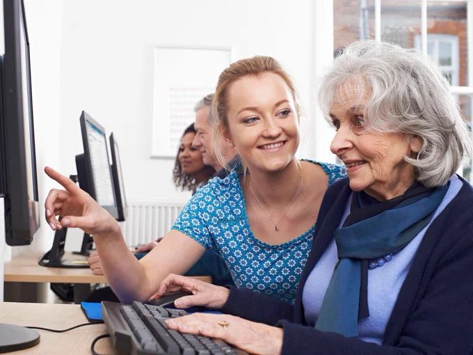 An image of a young individual crouching down and pointing at something on a computer screen, as they show an older adult how to use a mouse and keyboard during a class or workshop. Other students can be seen working on computers in the background. 