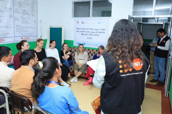 A group of people siting in a semi circle in a chair hearing a lady standing and facing them wearing a half jacket that reads UNFPA Nepal 