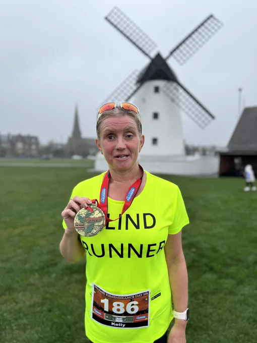 The image shows a woman standing on a grassy area in front of a large white windmill with black sails. She is wearing a bright yellow-green shirt with the words "BLIND RUNNER" printed in bold black letters on the front. Around her neck is a red lanyard holding a large medal, which she is holding up for display. The medal has an image of a windmill and the word "Windmill" on it. She is also wearing a race bib with the number 186 and the name "Kelly" on it. In the background, there is a tall, pointed structure, possibly a church spire, and some buildings. The sky is overcast.