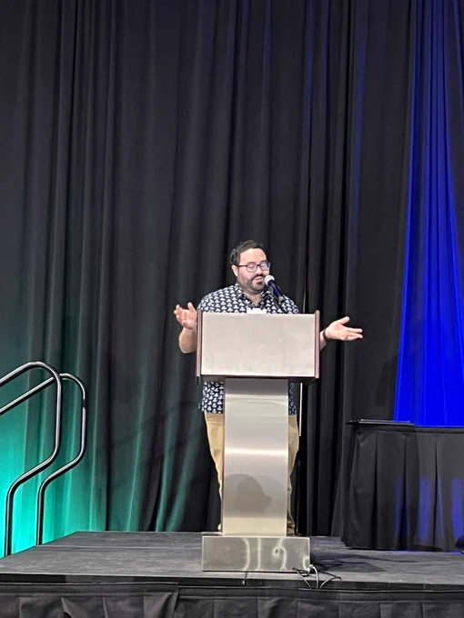 A man in glasses stands at a podium to give a speech before a crowded room while accepting an award.