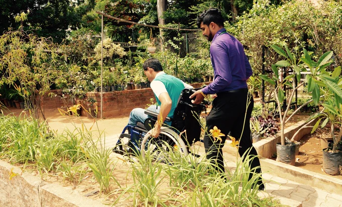 Man in a purple business shirt is pushing a man in wheelchair up a dirt path in a garden setting.