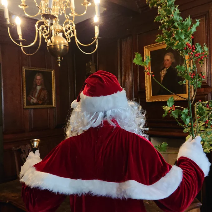 A person seen from the back wearing a red cloak and hat with white hair, holding holly and a handbell, standing in a room with dark wood pannelling and 2 paintings on the wall.
