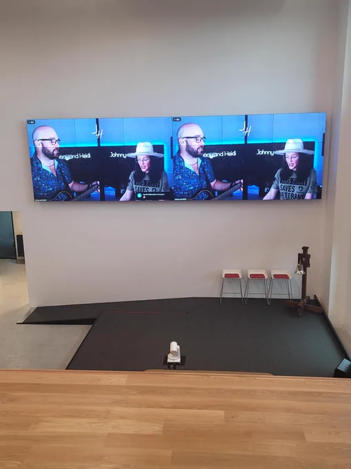 A room featuring a large television screen displaying three instances of the same image of a person wearing a white hat and glasses, playing a guitar. The screen is mounted on a white wall above a small stage area with a microphone stand. The name "Johnny" is visible on the screen.