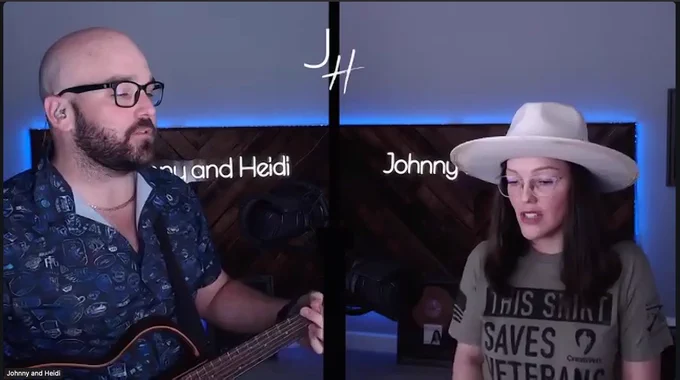 Johnny and Heidi sitting in a studio with their names displayed on the screen; Johnny holds a guitar, and Heidi wears a wide-brimmed hat, reading from a screen. The background features a wooden design with blue lighting.