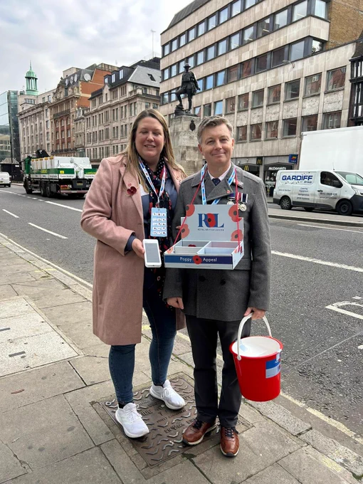 Two individuals wearing badges and holding a donation bucket for the Royal British Legion's Poppy Appeal stand on a city street.