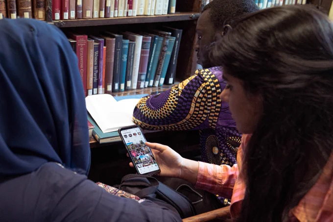 Three individuals studying together in a library with books and a smartphone on the table.