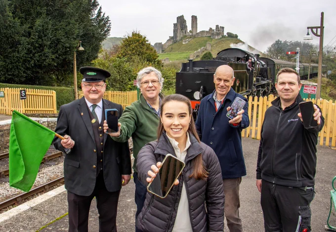 Five people on a railway platform with a steam training coming  into view. Four of them are holding out  smart phones, while one man in railway uniform is holding a green flag