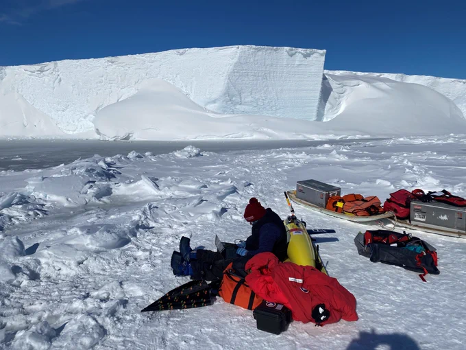 A scientist is sitting on the ice, with the ice shelf in the background. You can see the bright yellow seaglider, which the scientist is sitting against, with other kit on sleighs around them. 

Photo credit: Walker Smith