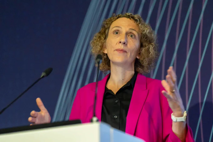 Stella Peace stood behind a lectern wearing a pink jacket and black shirt giving a speech with her hands up by her shoulders.    