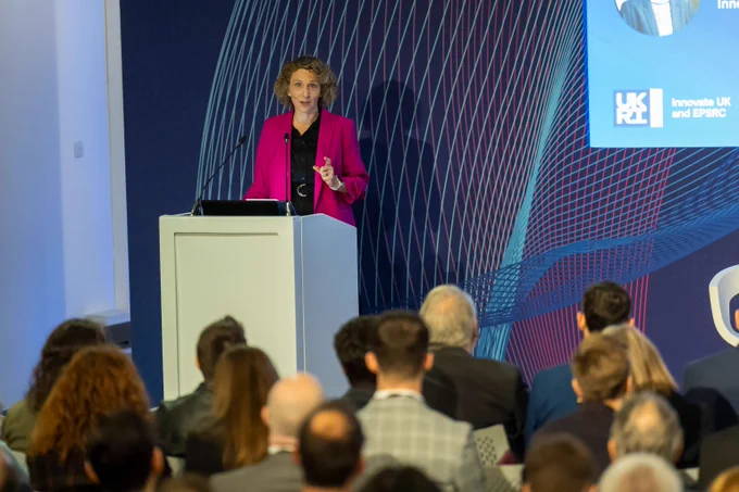 Stella Peace stood behind a lectern wearing a pink jacket and black shirt in front of a seated audience.   