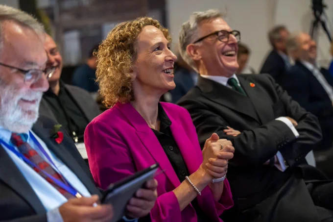 Stella Peace and Roger McKinlay in the audience both with smiles on their faces. Stella has her hands cupped together and Roger has his arms folded.   