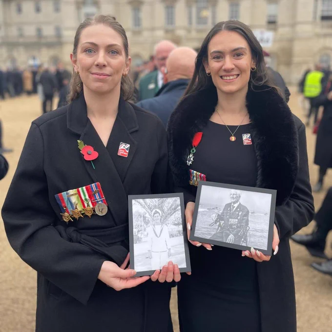George's granddaughters stand in front of the Horse Guards Parade in London,  each holding a framed black and white photograph of their late grandfather. One photo shows George in his naval uniform as a young soldier, and the other shows him later in life wearing his medals. Both wear poppies and his medals to honour his memory.