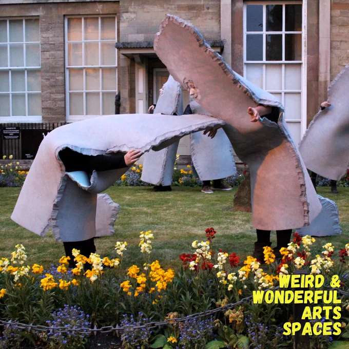 People wearing grey stone costumes with face cut-outs, standing on a grass patch with bright flower beds in front of them, and the Peterborough Museum behind them.